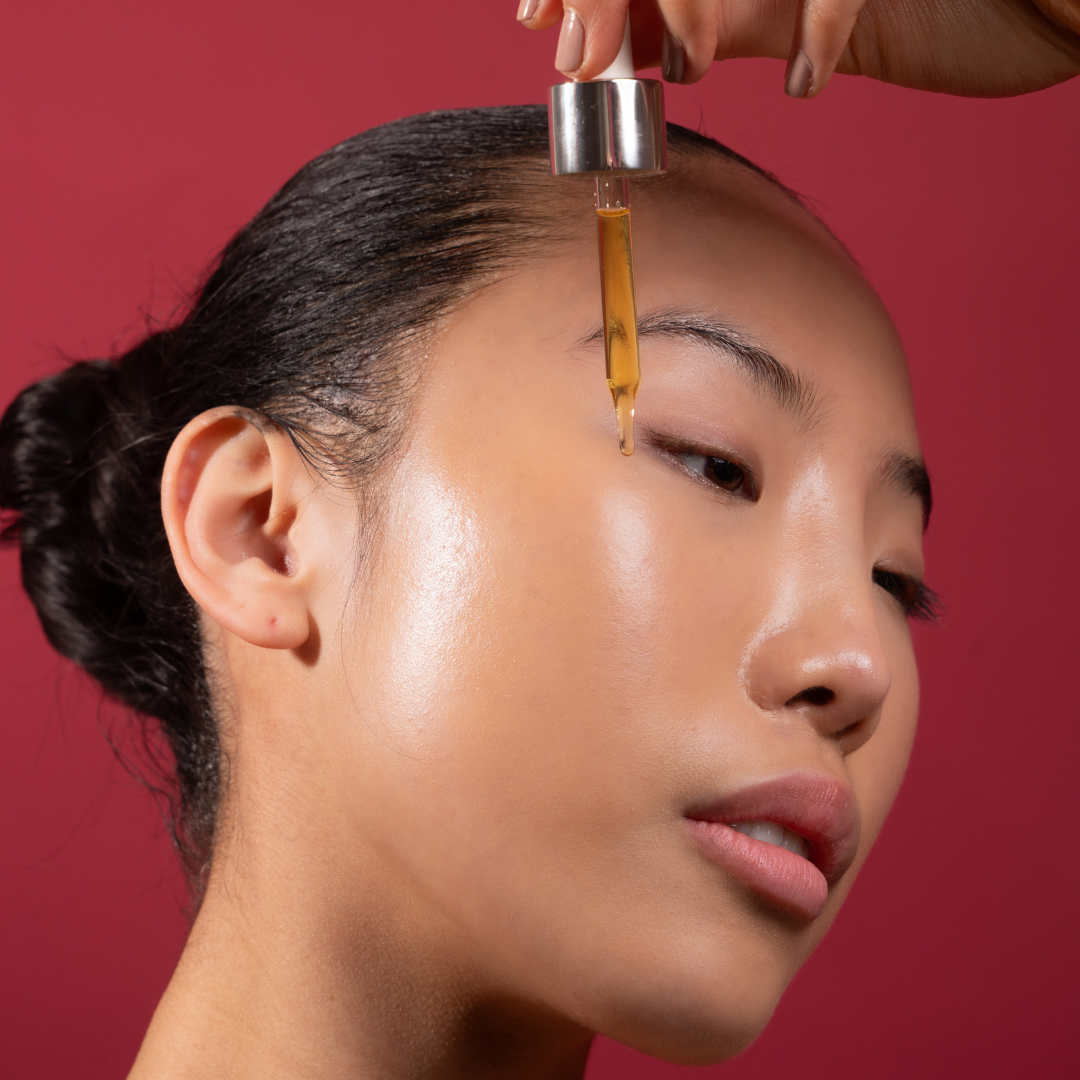 A close-up of a woman receiving a skincare treatment, with a hand holding a glass dropper above her forehead. The dropper contains a golden amber liquid, set against a solid dark red background.