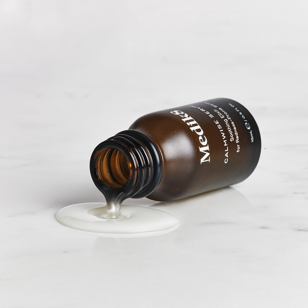 Open amber Medik8 Calmwise Serum bottle lying on its side with clear serum flowing onto a pale surface.
